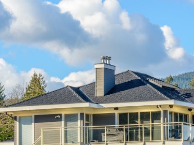 Modern house with dark shingle roof, gray chimney, and glass balcony railing, set against a partly cloudy blue sky with trees in the background.