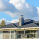 Modern house with dark shingle roof, gray chimney, and glass balcony railing, set against a partly cloudy blue sky with trees in the background.
